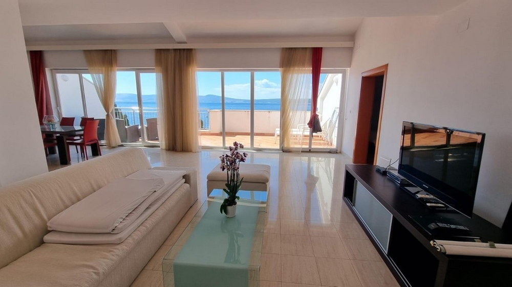 Widok na jasną kanapę, telewizor oraz morze i wybrzeże w tle.