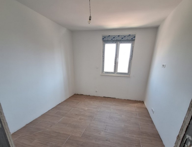 Pusty pokój w stanie surowym nowego mieszkania z drewnianą podłogą i oknem od frontu, białymi ścianami, podłączeniem oświetlenia sufitowego, posiadłość Panorama Scouting w Chorwacji.