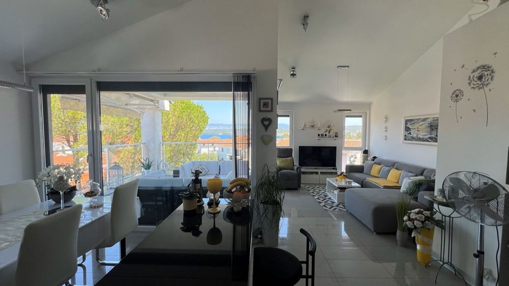 Jasny salon z szarą sofą, jadalnią i dużym oknem wychodzącym na taras z widokiem na morze.