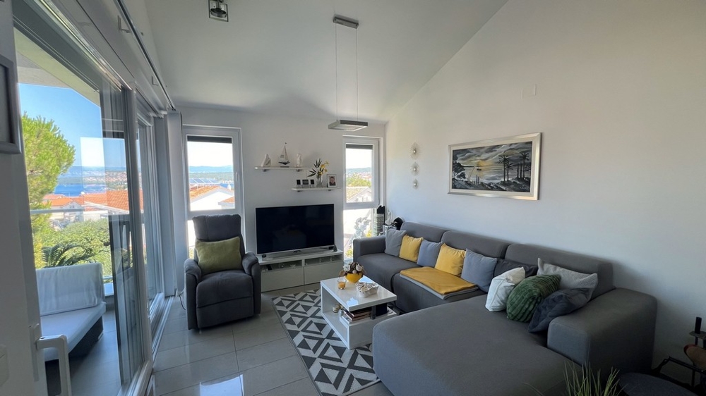 Nowoczesny salon z dużym telewizorem, szarą sofą i widokiem na morze przez duże okna.