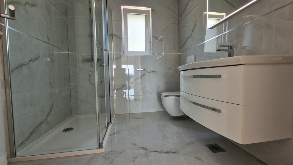 Nowoczesna łazienka z prysznicem, toaletą i toaletką, jasne płytki i okna.