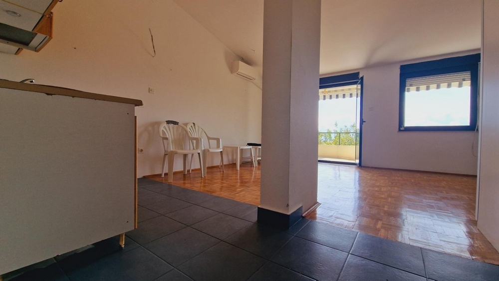 Pokój dzienny z otwartym przejściem do kuchni, prostymi białymi meblami i podłogą wyłożoną kafelkami.