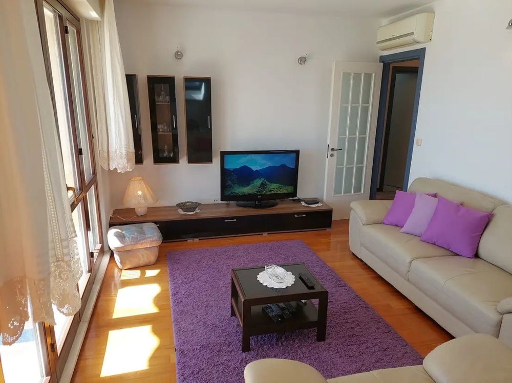 Pokój dzienny z sofą, telewizorem, fioletowymi poduszkami i dywanem, dużymi oknami