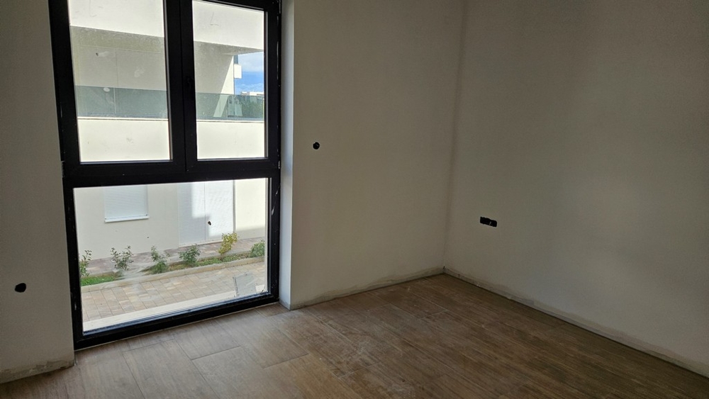 Kolejny pokój z dużymi oknami, widok na nowoczesny budynek i teren utwardzony.