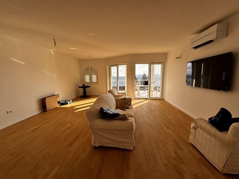 Jasny salon z białą sofą, telewizorem naściennym i panoramicznymi oknami z widokiem na morze