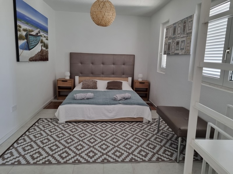 Na sprzedaż sypialnia w apartamentowcu w pierwszym rzędzie od morza w Karlobag w Chorwacji, z dużym podwójnym łóżkiem, stolikami nocnymi i obrazem przedstawiającym łódź na morzu.