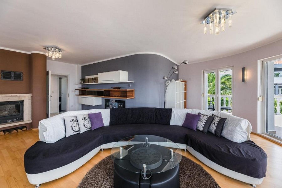 Elegancki salon w chorwackim apartamentowcu z widokiem na morze, z dużą zakrzywioną sofą i okrągłym szklanym stołem na brązowym dywanie, kominkiem i otwartym balkonem.