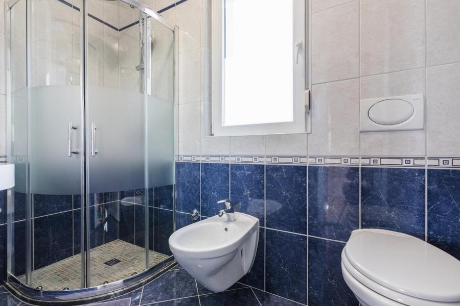Łazienka w nieruchomości w Chorwacji, apartamentowiec z widokiem na morze, z narożnym prysznicem, bidetem i toaletą, jasne ściany i ciemnoniebieskie płytki