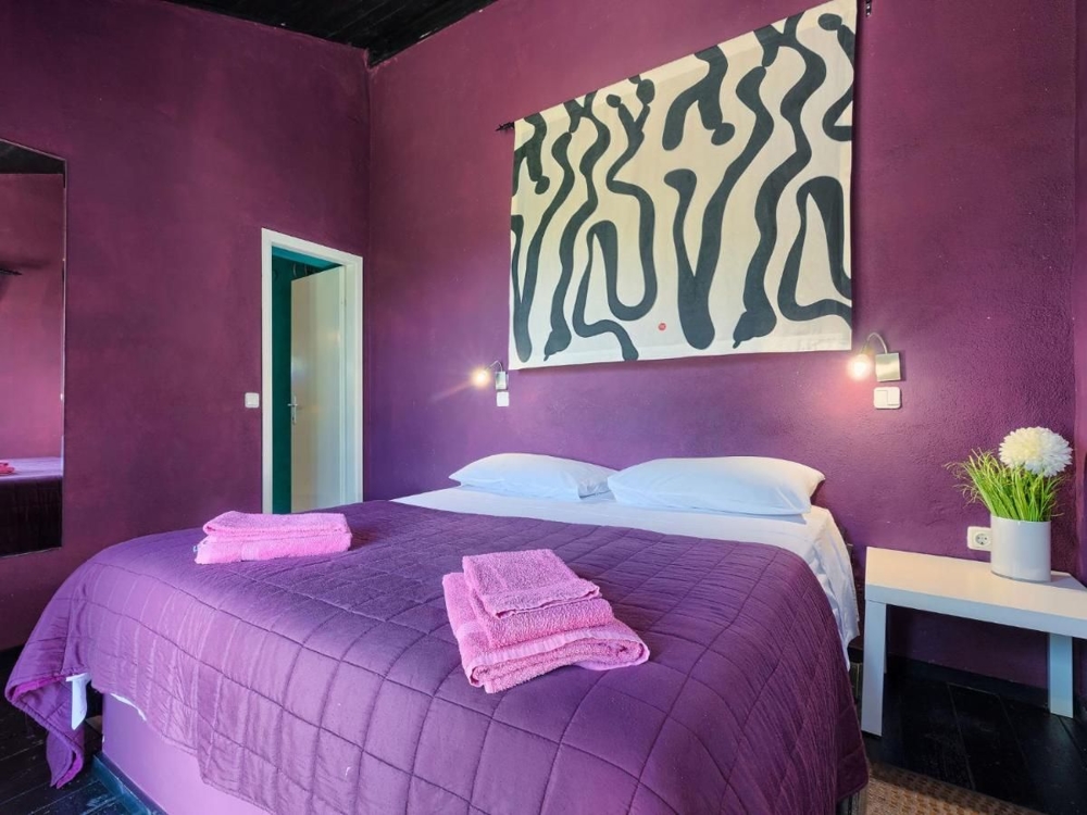 Sypialnia w nieruchomości w Chorwacji, z fioletowymi ścianami, dużym obrazem z abstrakcyjnymi wzorami, dwiema lampkami nocnymi i pościelą w dopasowanym fioletowym odcieniu