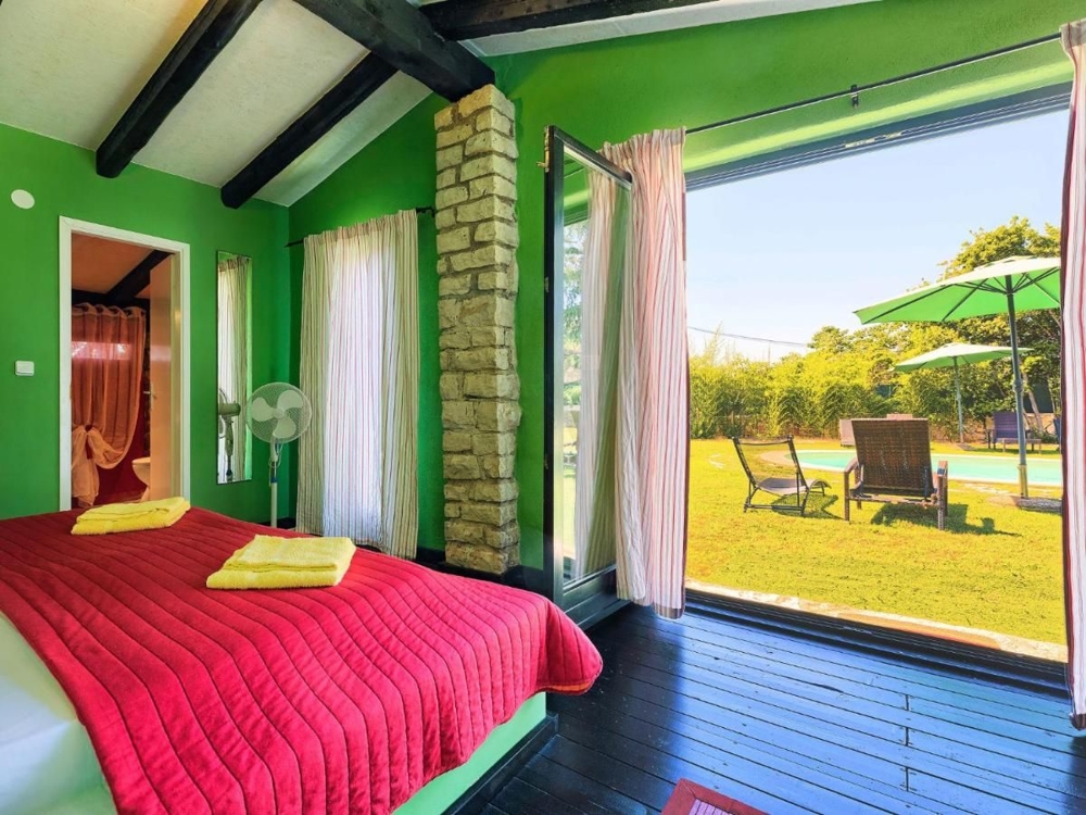 Jasna sypialnia z widokiem na ogród, część nieruchomości w Chorwacji, z zielonymi ścianami, czerwoną narzutą i wyjściem na taras z leżakami i parasolem