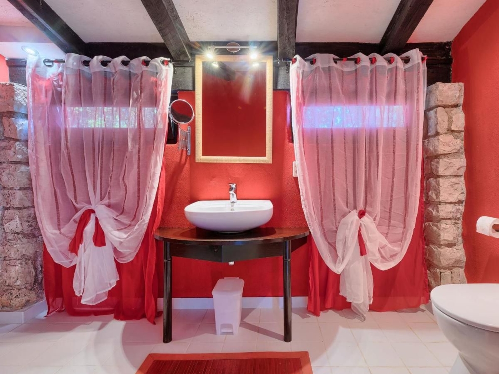 Łazienka nieruchomości w Chorwacji z czerwonymi ścianami, białą instalacją wodną, ​​zlewem na rustykalnym stole i przezroczystymi czerwonymi zasłonami w oknach