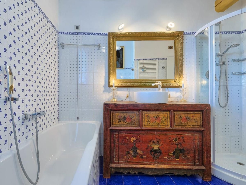 Łazienka w chorwackiej posiadłości ozdobiona niebiesko-białymi kafelkami, wanną, kabiną prysznicową i zabytkową kolorową szafką pod złotym lustrem
