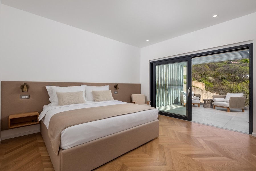 Minimalistyczna sypialnia z dużym podwójnym łóżkiem, beżową pościelą i zagłówkiem, drewnianą podłogą i przesuwanymi drzwiami prowadzącymi na taras z częścią wypoczynkową i pięknym widokiem.