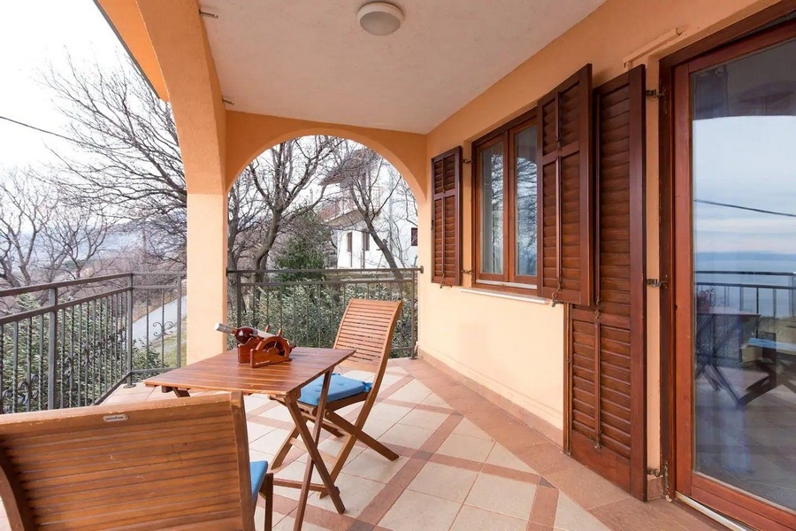 Zadaszony balkon z drewnianymi krzesłami i stołem, z widokiem na zieloną okolicę i morze w oddali.