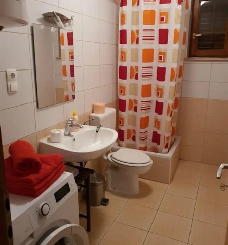 Funkcjonalna łazienka z zasłoną prysznicową, pralką oraz dodatkami w kolorze czerwono-pomarańczowym, wyposażona w umywalkę, toaletę i małe okienko.