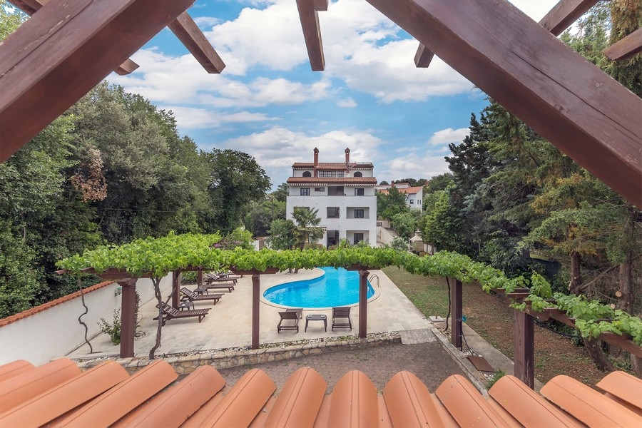 Widok z pergoli na strefę basenową i biały apartamentowiec otoczony bujną roślinnością