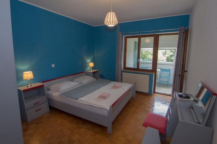 Sypialnia ze ścianami pomalowanymi na niebiesko, dużym podwójnym łóżkiem, dwiema szafkami nocnymi z lampkami, oknem i drzwiami prowadzącymi na balkon.