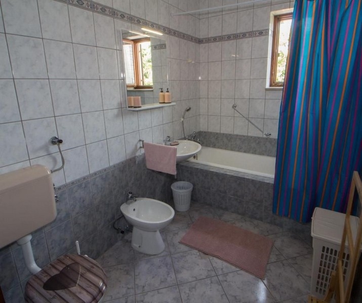 Łazienka z biało-szarymi płytkami, wanną, umywalką, bidetem, toaletą i niebieską zasłoną prysznicową. Małe okna wpuszczają naturalne światło.