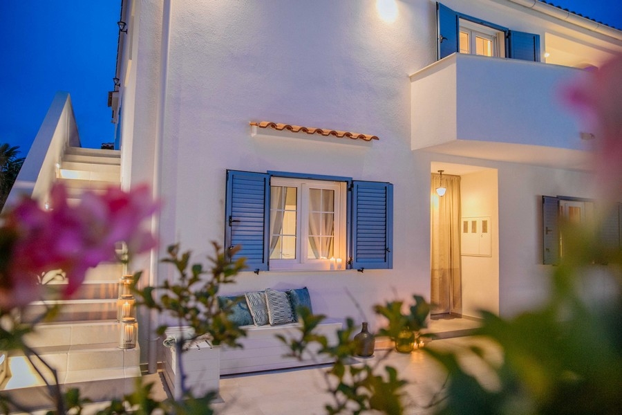 Widok z przodu śródziemnomorskiego domu z niebieskimi okiennicami i częścią wypoczynkową na świeżym powietrzu o zmierzchu, oświetlonym miękkim światłem