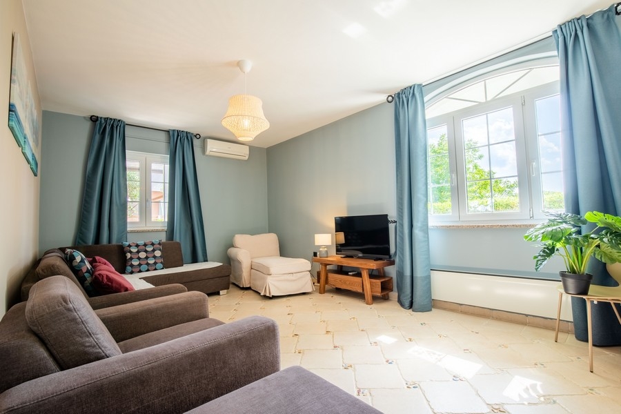 Przytulny salon z kilkoma sofami, telewizorem i dużymi oknami z niebieskimi zasłonami, które wpuszczają naturalne światło