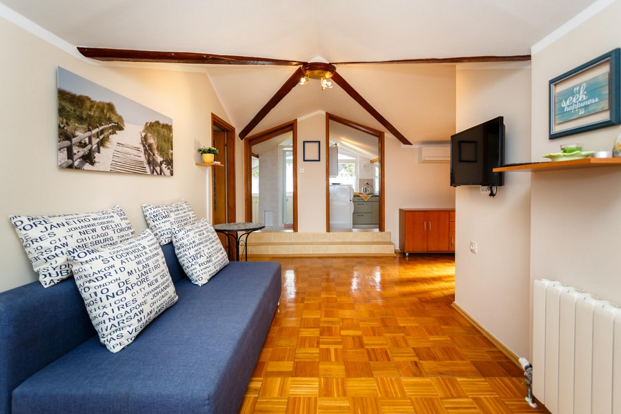 Pokój dzienny z sofą, drewnianą podłogą i belkami stropowymi prowadzi do małej kuchni