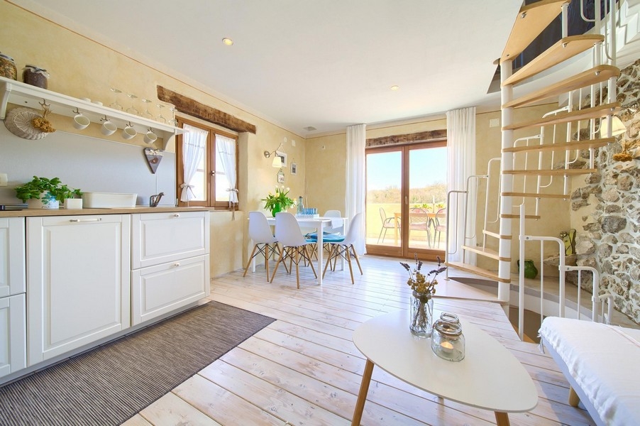 Otwarta część dzienna z nowoczesną kuchnią, stołem jadalnym i wyjściem na taras, ozdobiona jasnymi meblami.