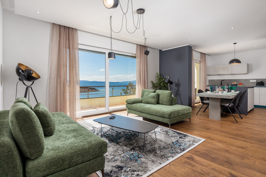 Salon z dużymi oknami, stylowymi meblami i widokiem na morze, w tle otwarta kuchnia.