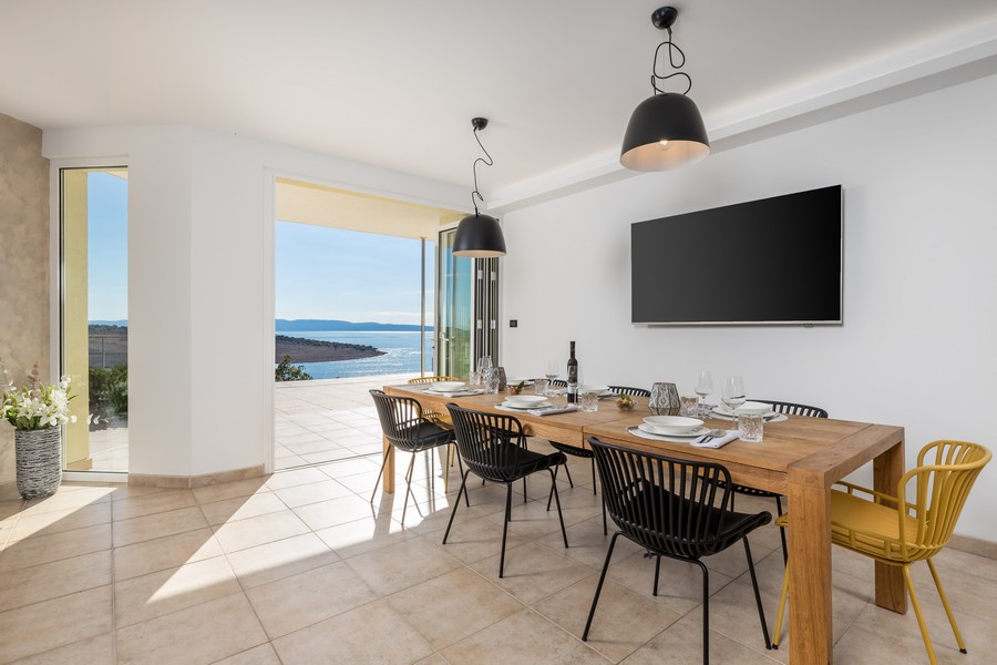 Jadalnia z dużym drewnianym stołem, nowoczesnymi krzesłami i bezpośrednim dostępem do tarasu z widokiem na morze.