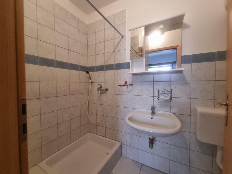 Jasna łazienka z płytkami, prysznicem i prostą umywalką.