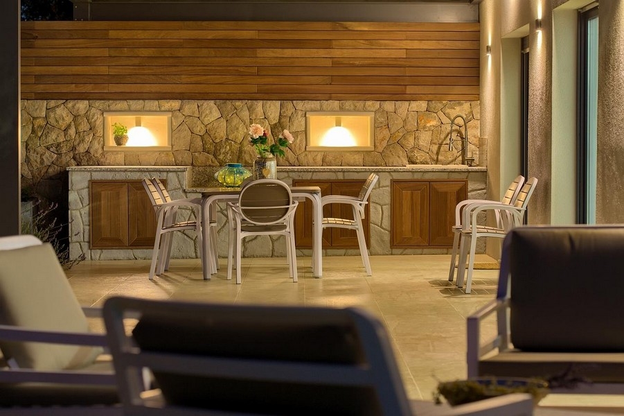 Zadaszona jadalnia na świeżym powietrzu z kamiennym aneksem kuchennym i drewnianymi szafkami, stylowo oświetlona