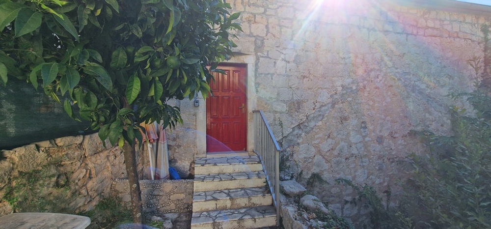 Mały obszar na zewnątrz z kamiennymi schodami prowadzącymi do czerwonych drewnianych drzwi. Ściana domu wykonana jest z kamienia naturalnego, a na niebie świeci słońce.