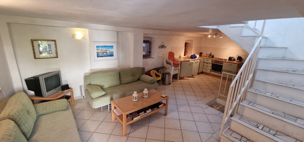 Przytulny salon na otwartym planie z zielonymi sofami, stolikiem kawowym, telewizorem i zintegrowaną kuchnią z tyłu. Podłogę pokrywają jasne płytki.