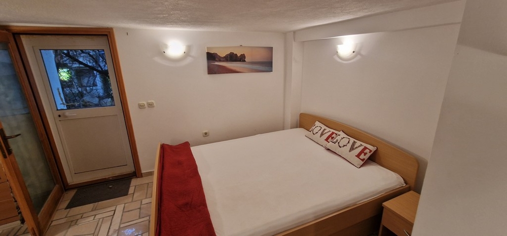 Sypialnia z łóżkiem małżeńskim ozdobiona białymi i czerwonymi akcentami. Na ścianie wisi zdjęcie plaży i drzwi prowadzące na zewnątrz.